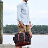 A man in a light shirt and khaki shorts carries a Tom Beckbe Canvas Carryall Bag by the water with trees behind.