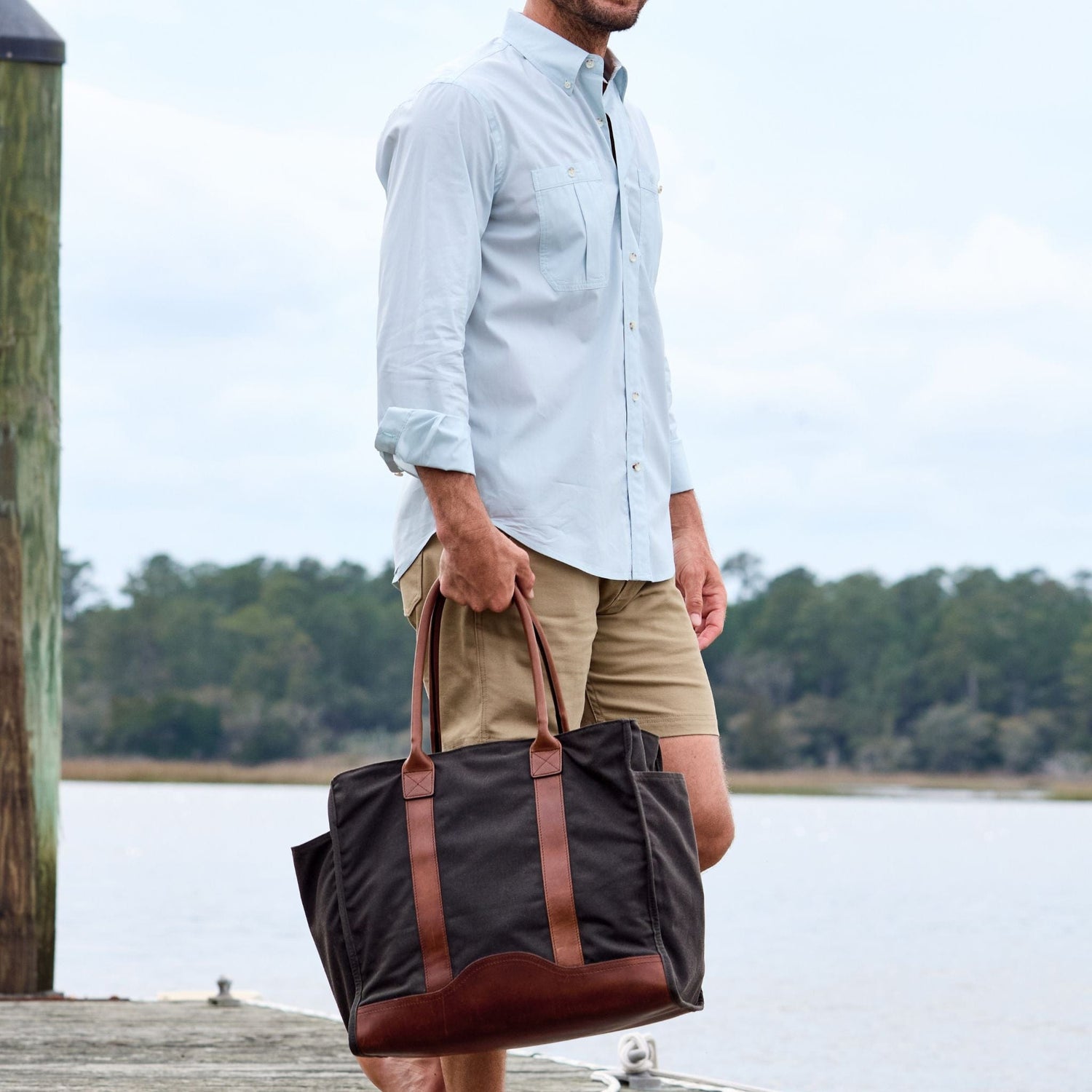 A man in a light shirt and khaki shorts carries a Tom Beckbe Canvas Carryall Bag by the water with trees behind.