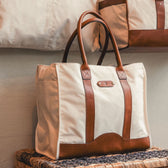 The Tom Beckbe Perdido Canvas Carryall Bag in beige with brown leather handles rests on a woven surface.