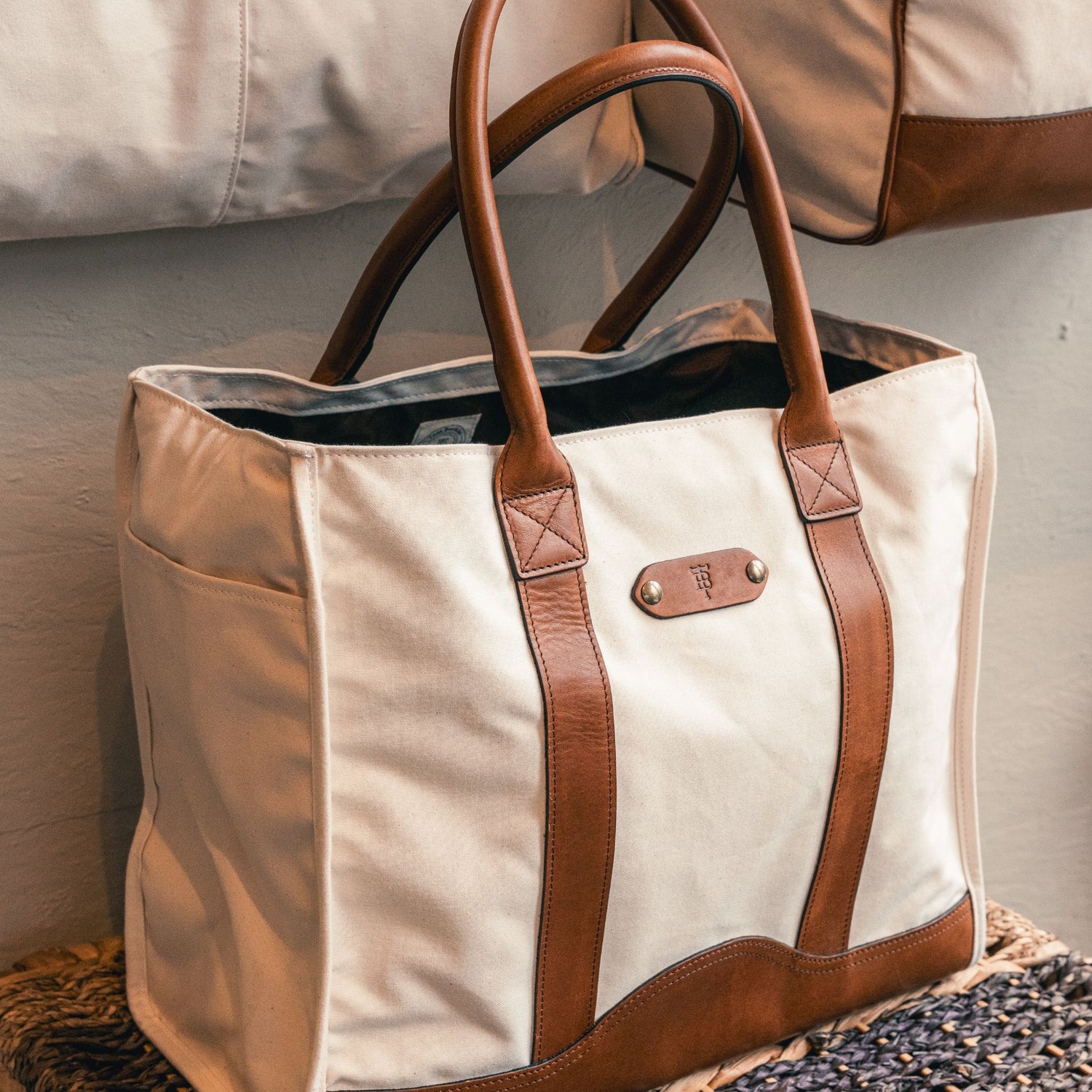The Perdido Canvas Carryall Bag by Tom Beckbe features beige canvas and brown leather handles on a woven surface.