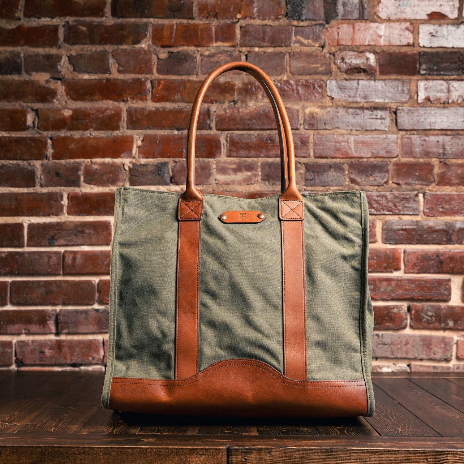 Tom Beckbes Canvas Carryall Bag in waxed cotton, with brown leather handles, sits on wood next to a brick wall.