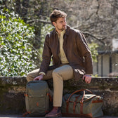 A man sits on a stone wall outdoors in a brown jacket and khaki pants, next to two Tom Beckbe Canvas Daypacks.