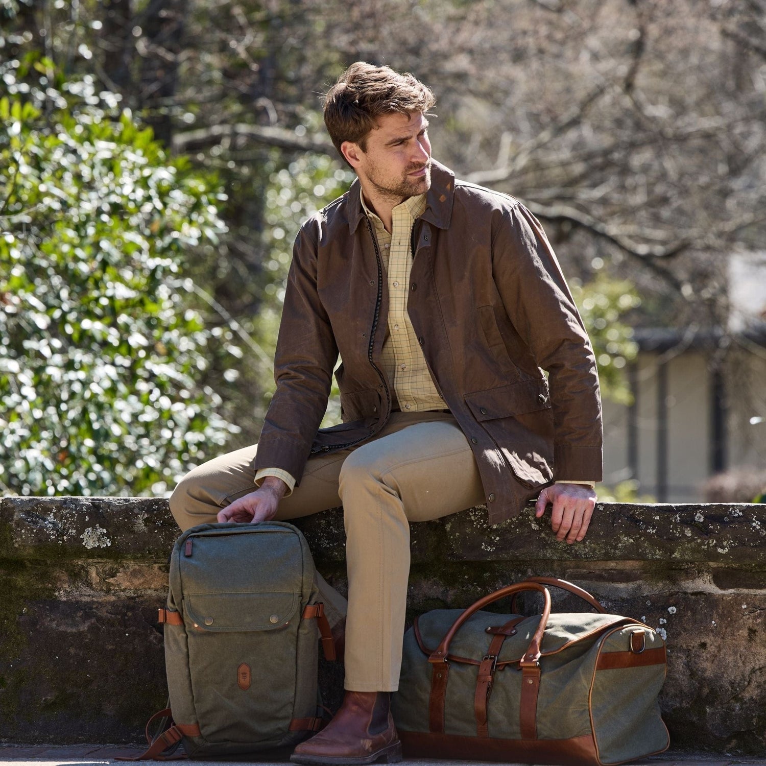 A man sits on a stone wall outdoors in a brown jacket and khaki pants, next to two Tom Beckbe Canvas Daypacks.