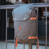 Tom Beckbes Canvas Daypack in gray with brown straps hangs on a railing, blurred storefront behind.