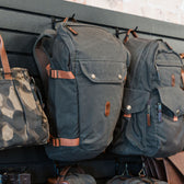 Three Tom Beckbe Canvas Daypacks with adjustable straps and padded laptop sleeves are on display on a black rack.