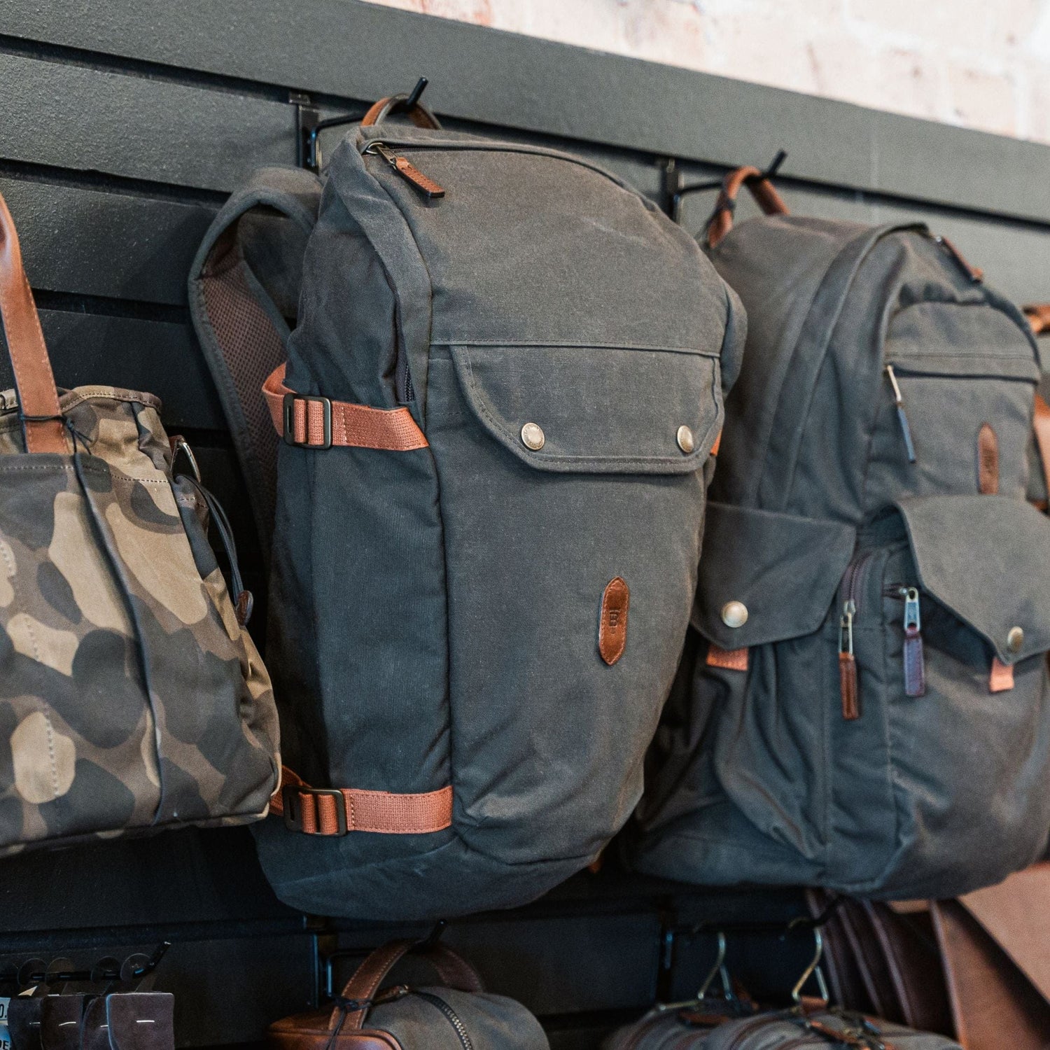 Three Tom Beckbe Canvas Daypacks with adjustable straps and padded laptop sleeves are on display on a black rack.