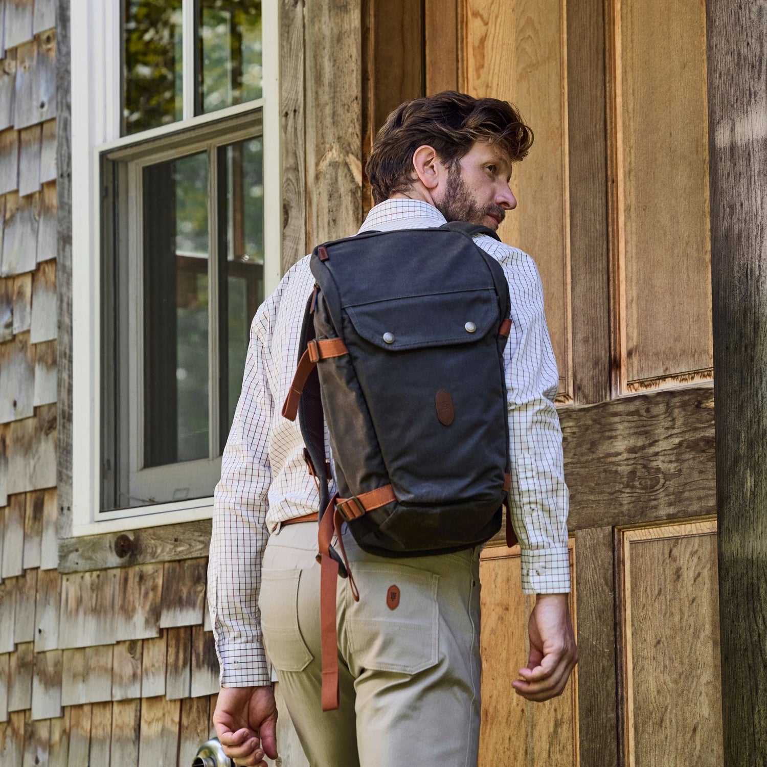 Man with short hair, in a checkered shirt and beige pants, carrying a Tom Beckbe Canvas Daypack by a wooden building.