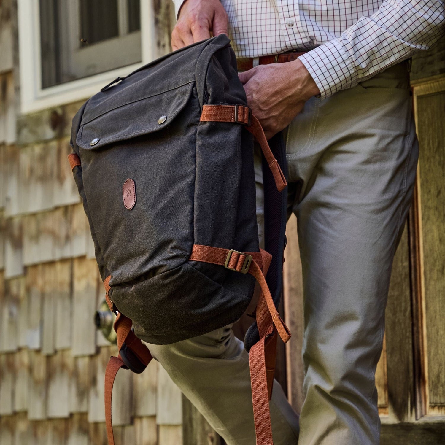 Person holding a Tom Beckbe Canvas Daypack with brown straps, standing in front of a wooden wall and door.