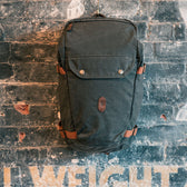 Tom Beckbes dark green Canvas Daypack with leather straps hangs on a textured brick wall with faded lettering.