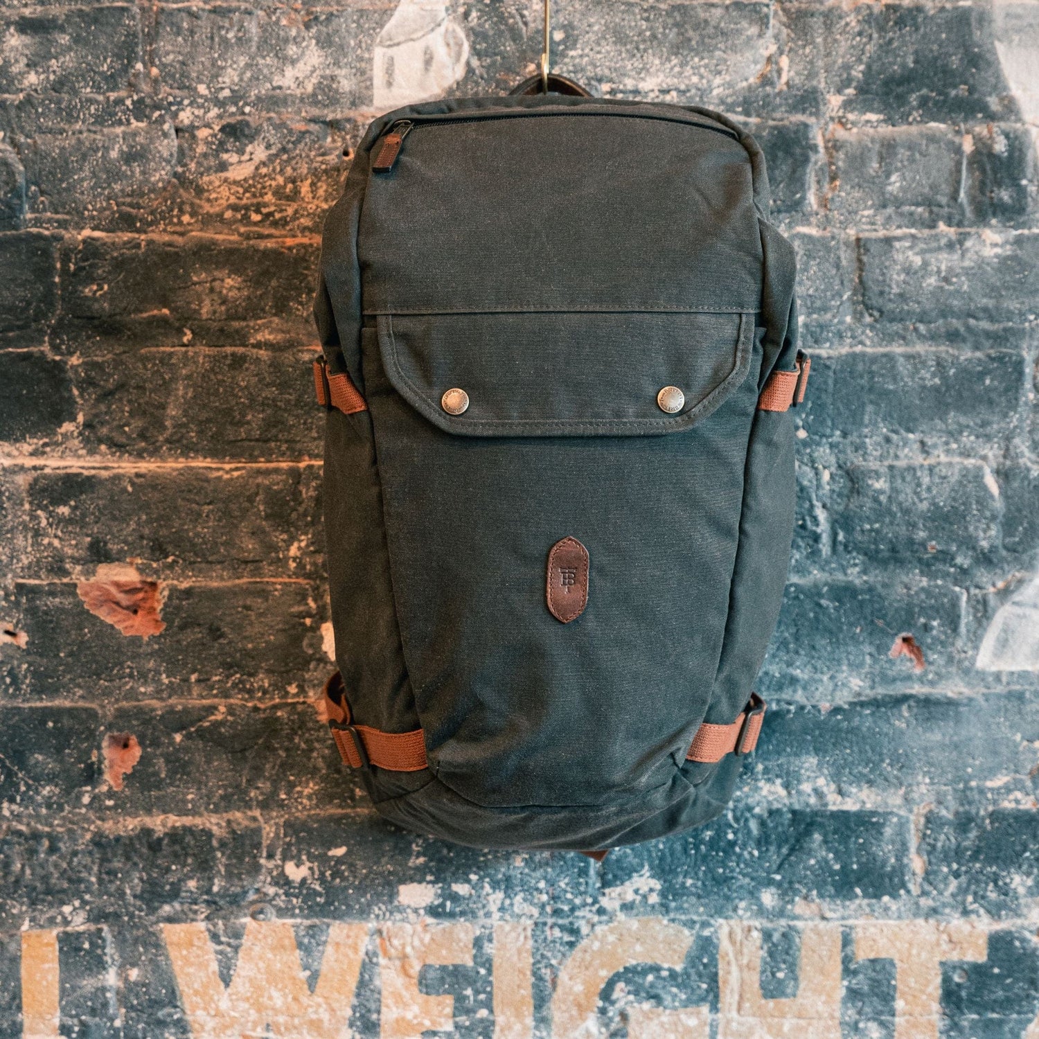Tom Beckbes dark green Canvas Daypack with leather straps hangs on a textured brick wall with faded lettering.