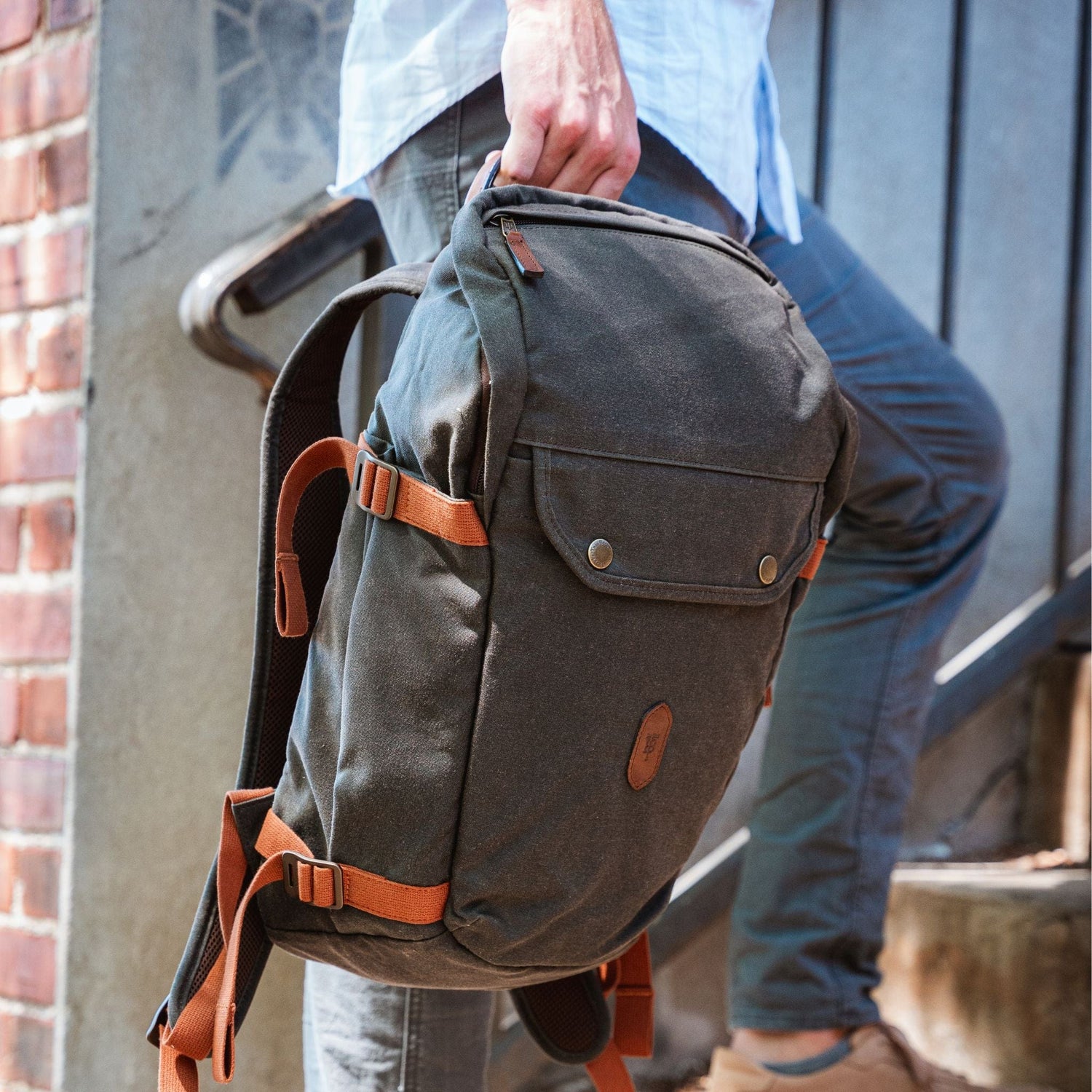 A person with a Tom Beckbe Canvas Daypack, featuring adjustable shoulder straps, walks up steps beside a brick wall.