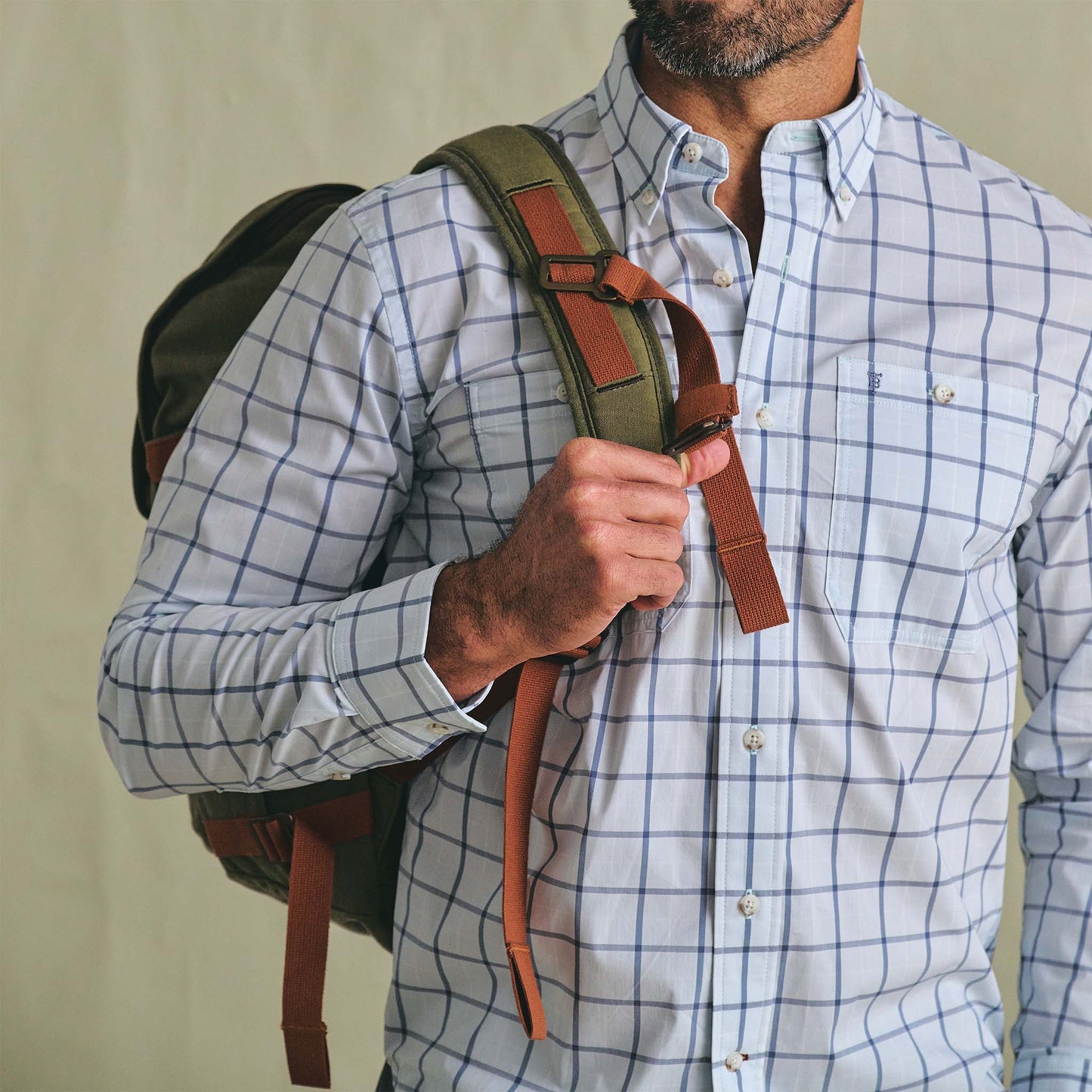 A man in a blue checked shirt carries his Tom Beckbe Canvas Daypack over one shoulder, holding the strap with one hand.