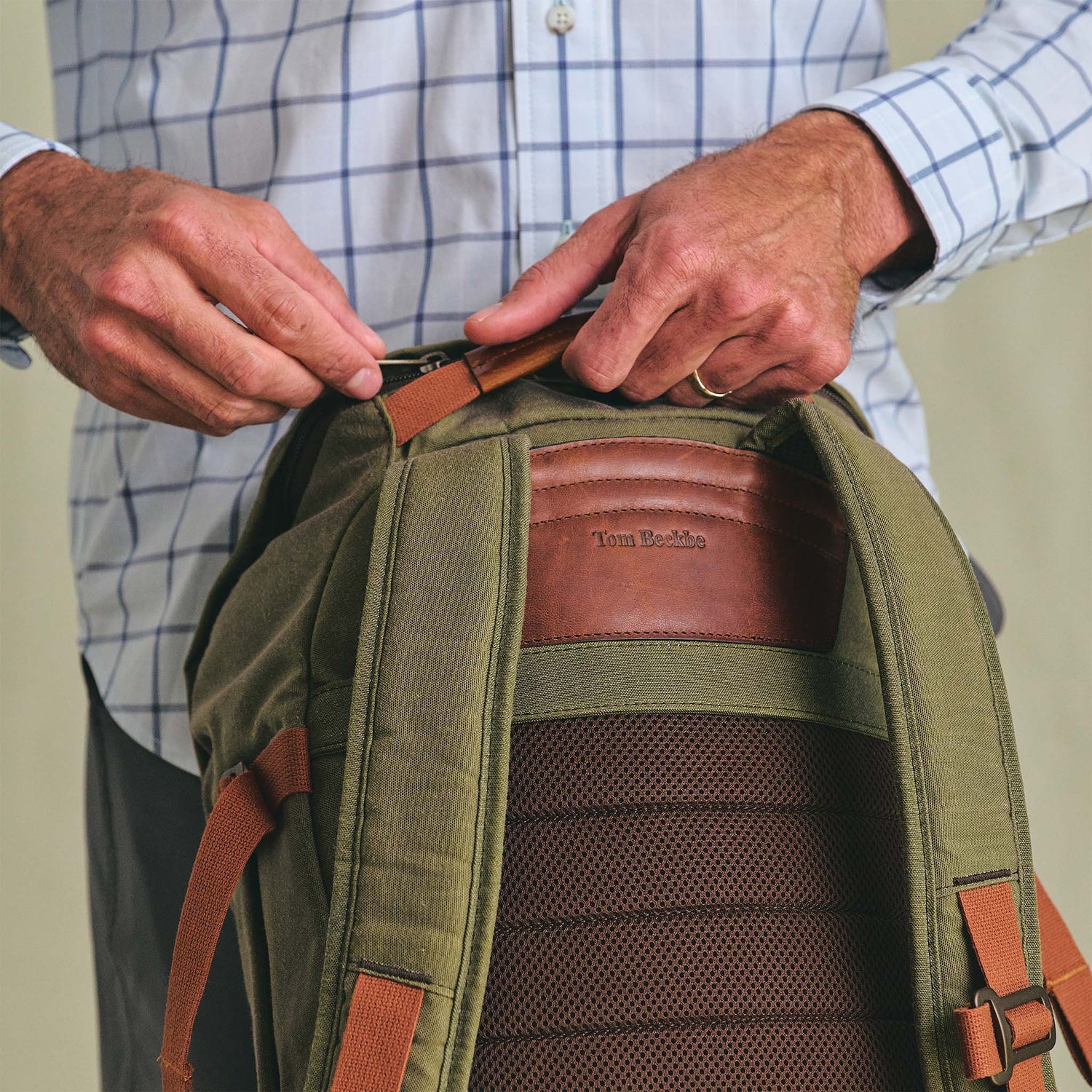 Person in a plaid shirt unzipping an olive green Tom Beckbe Canvas Daypack with a leather name patch.