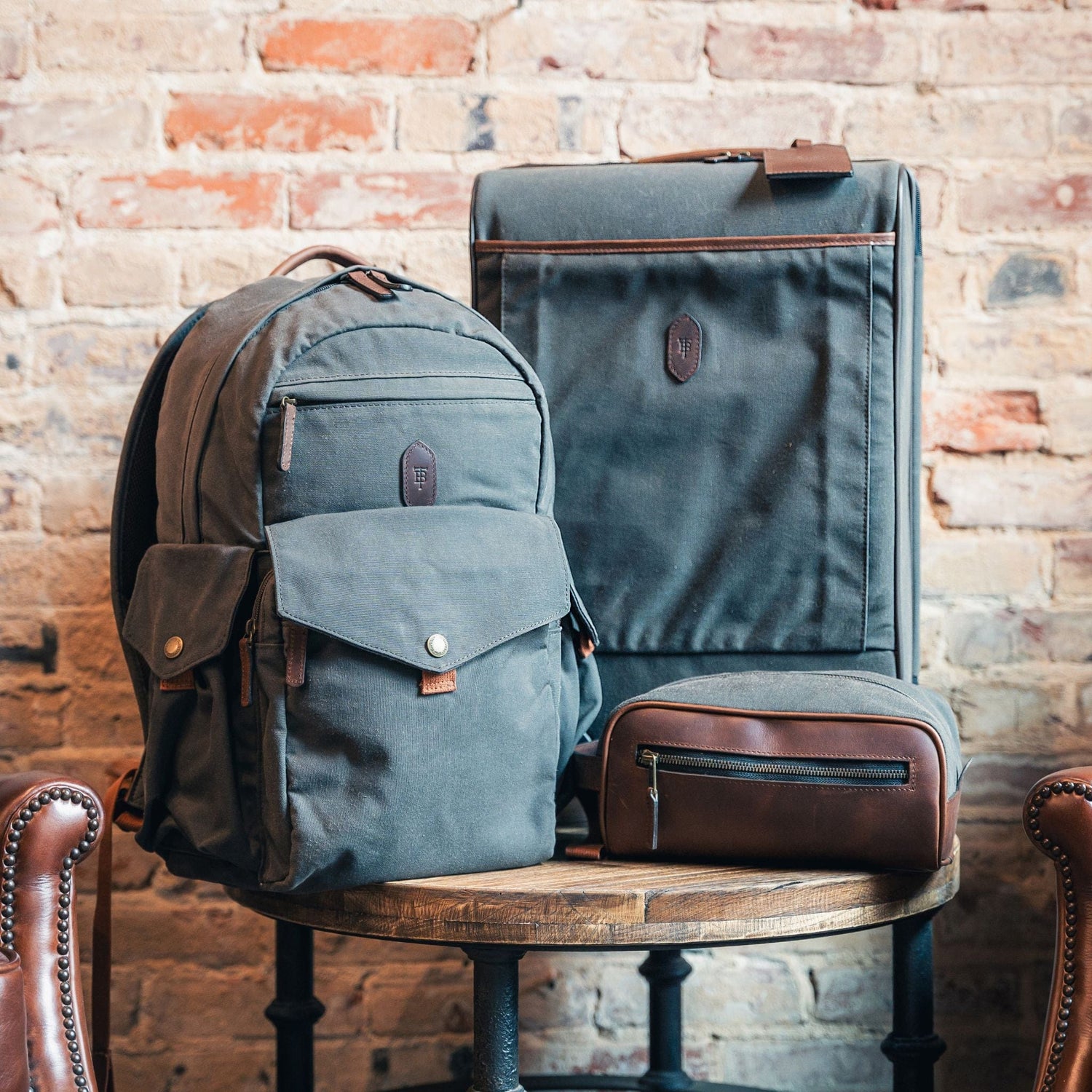 A gray Canvas Dopp Kit by Tom Beckbe sits with a backpack and suitcase on wood against a rustic brick wall.