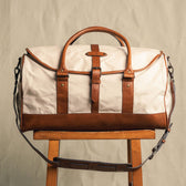 The Tom Beckbe Perdido Canvas Weekender Duffel Bag in beige and brown rests on a wooden stool against a plain background.