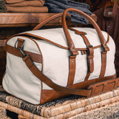 The Tom Beckbe Perdido Canvas Weekender Duffel Bag in beige with brown leather straps rests on a woven basket.