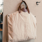 The Perdido Canvas Garment Bag by Tom Beckbe features brown leather handles and hangs from a wall-mounted hook.