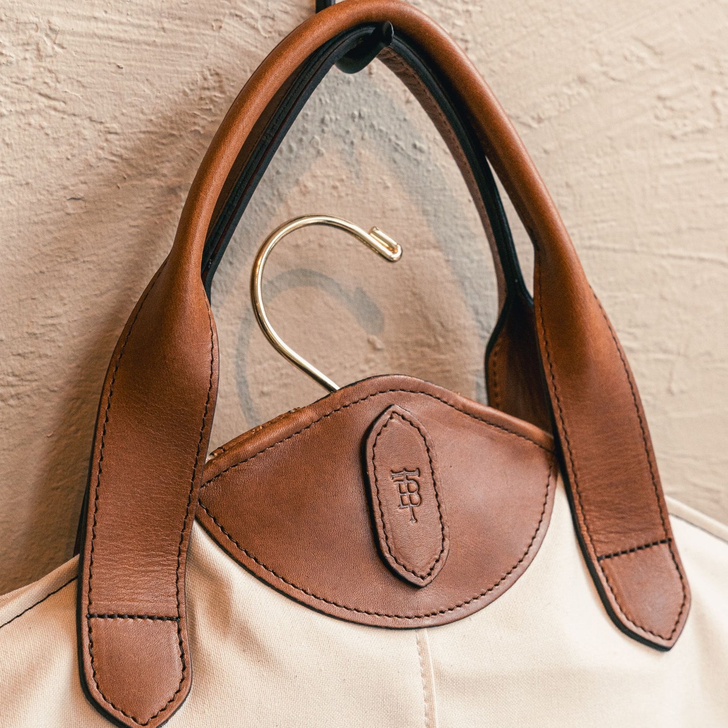 The Tom Beckbe Perdido Canvas Garment Bag in beige with brown leather handles hangs on a gold hook by a textured wall.
