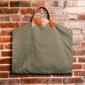Tom Beckbes Canvas Garment Bag with waterproof nylon and leather handles hangs against a brick wall.