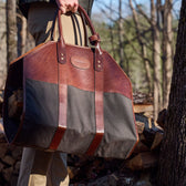 A person carries a Tom Beckbe Canvas Log Carrier filled with logs among stacked wood and trees.