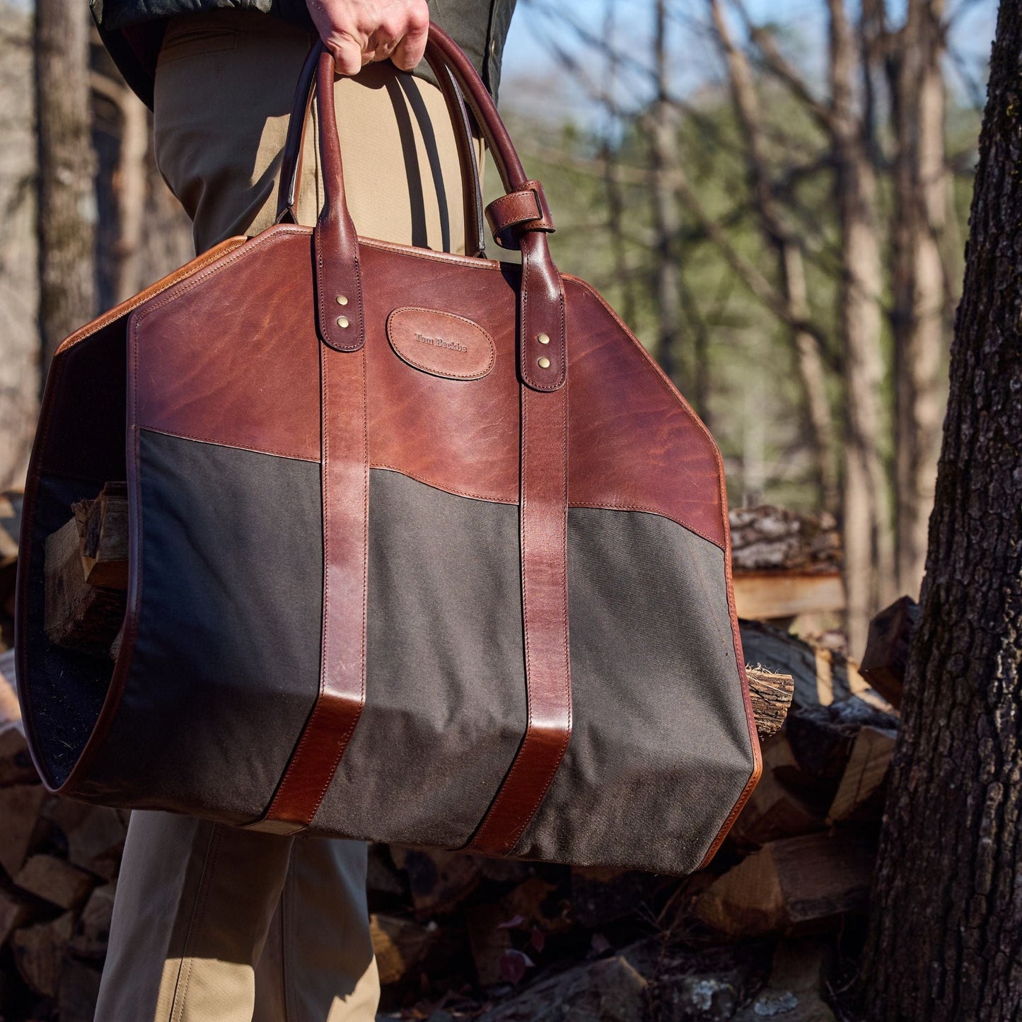A person carries a Tom Beckbe Canvas Log Carrier filled with logs among stacked wood and trees.