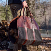 A person holds a Tom Beckbe Canvas Log Carrier filled with logs, standing by a woodpile near a lake.