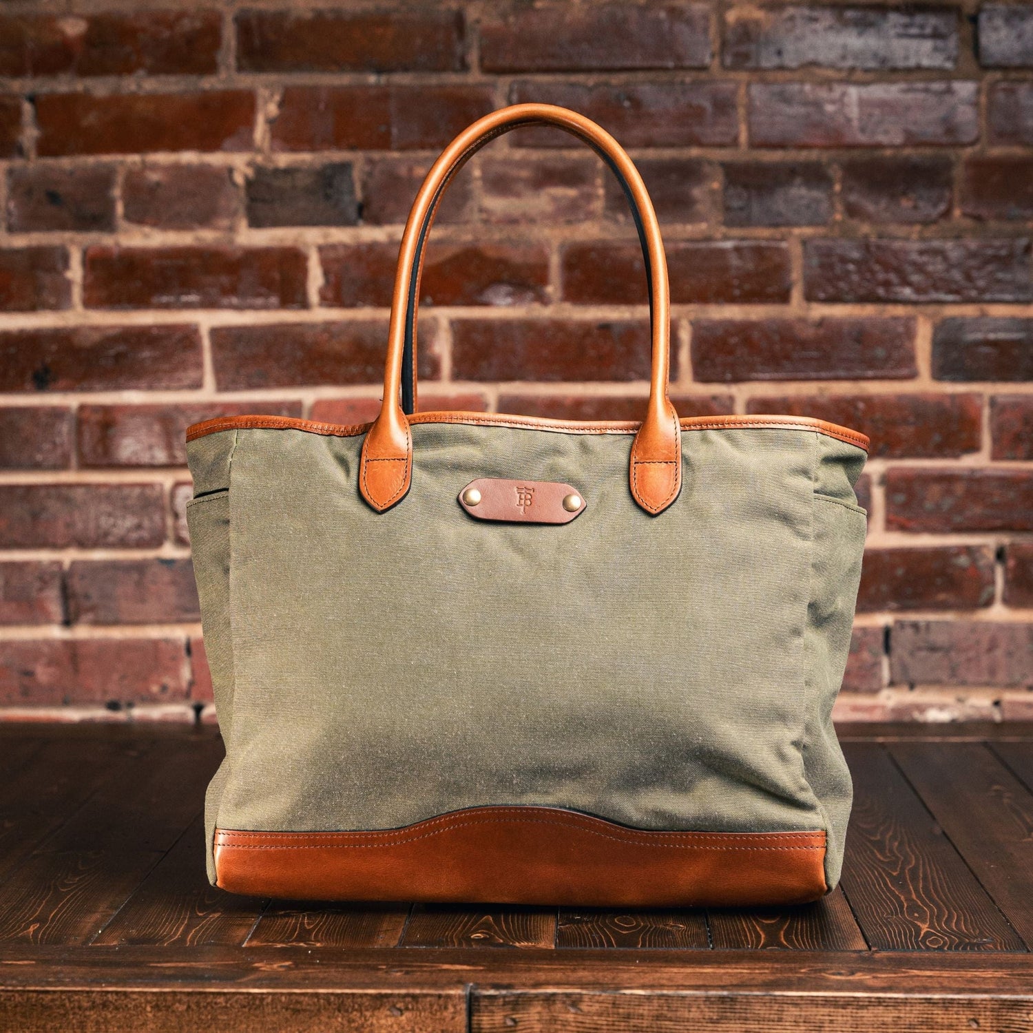 Tom Beckbes Canvas Tote in durable green and brown leather rests on a wooden surface against a brick wall.