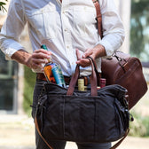 A person with a black Tom Beckbe Canvas Tailgater Bag full of bottles and whiskey, carrying a brown leather bag.