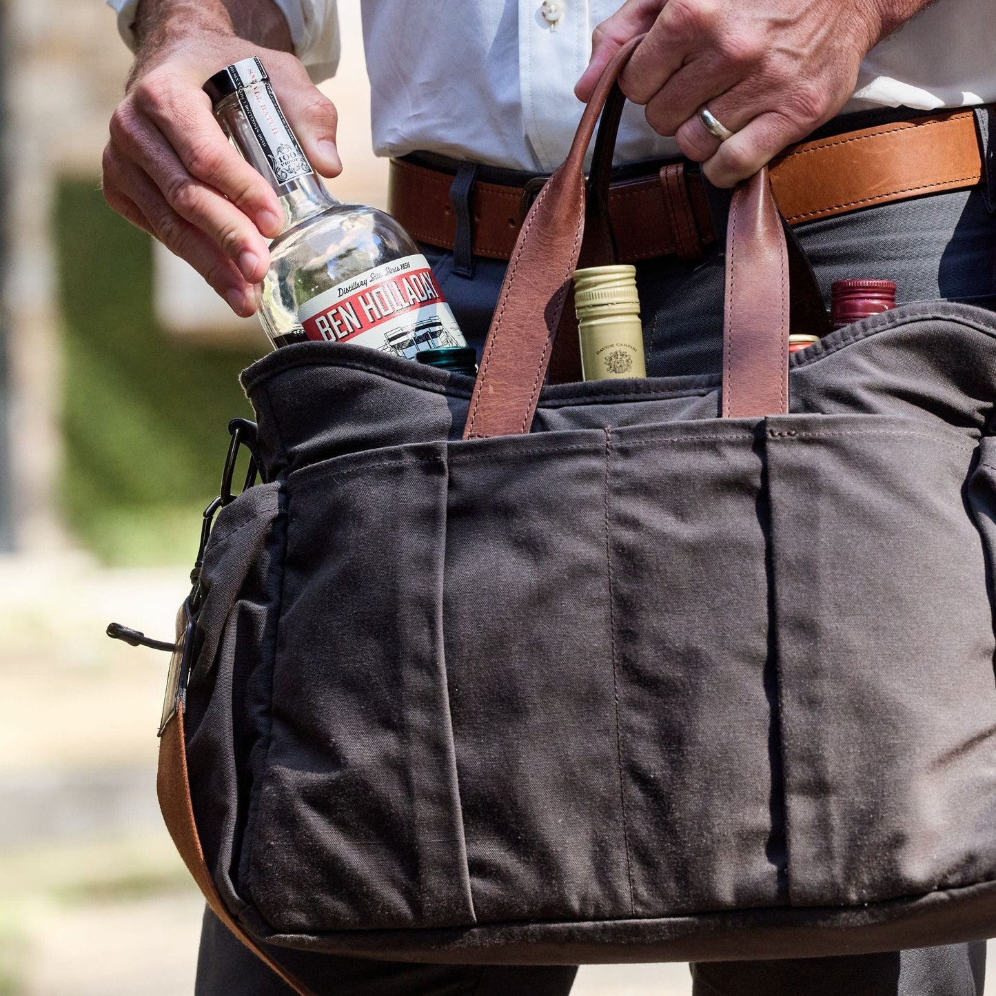 A person slips a Ben Holladay bottle into the pocket of a Tom Beckbe Canvas Tailgater Bag outdoors.
