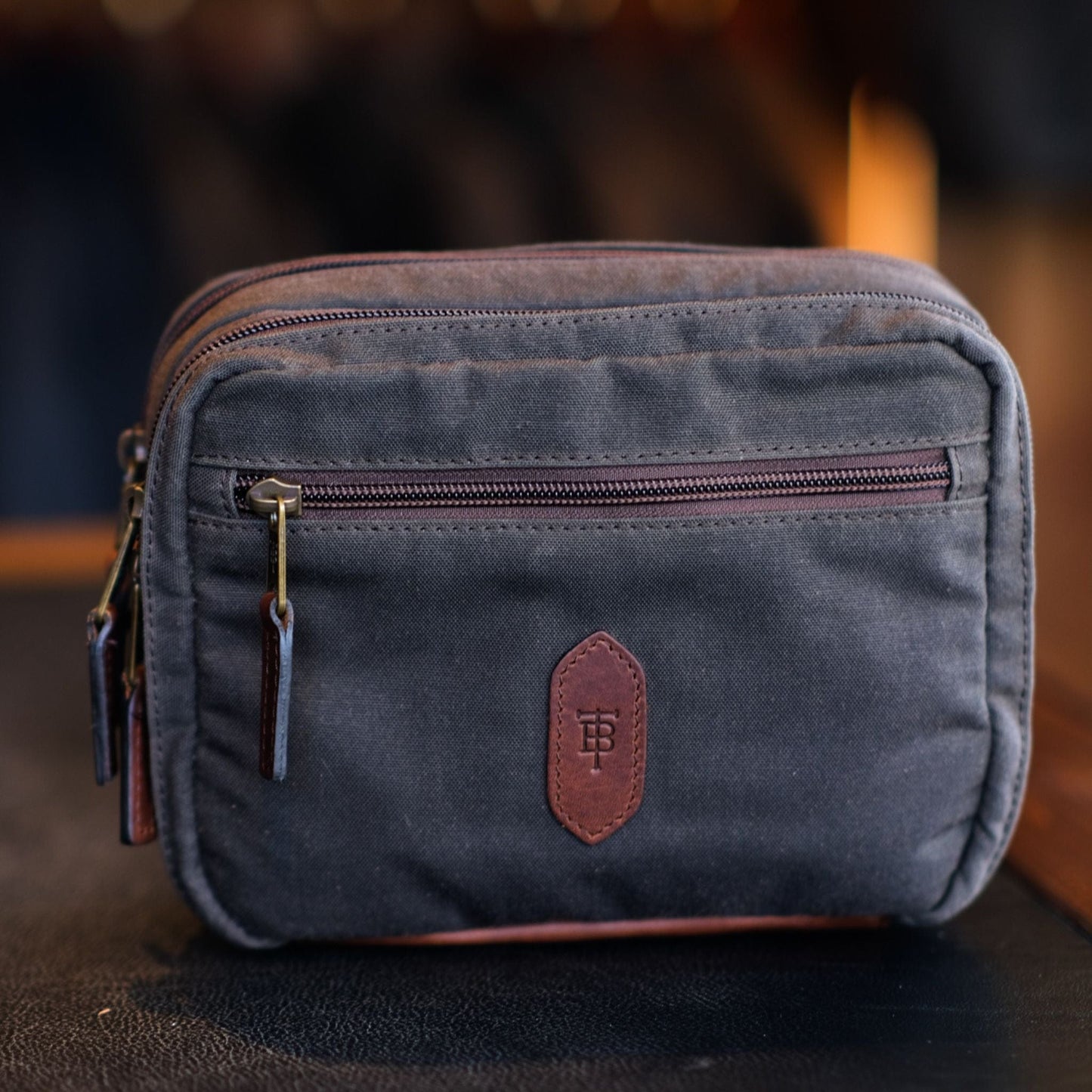 A Tom Beckbe dark gray Canvas Hanging Toiletry Bag with leather trim and a front zip pocket on a blurred background.