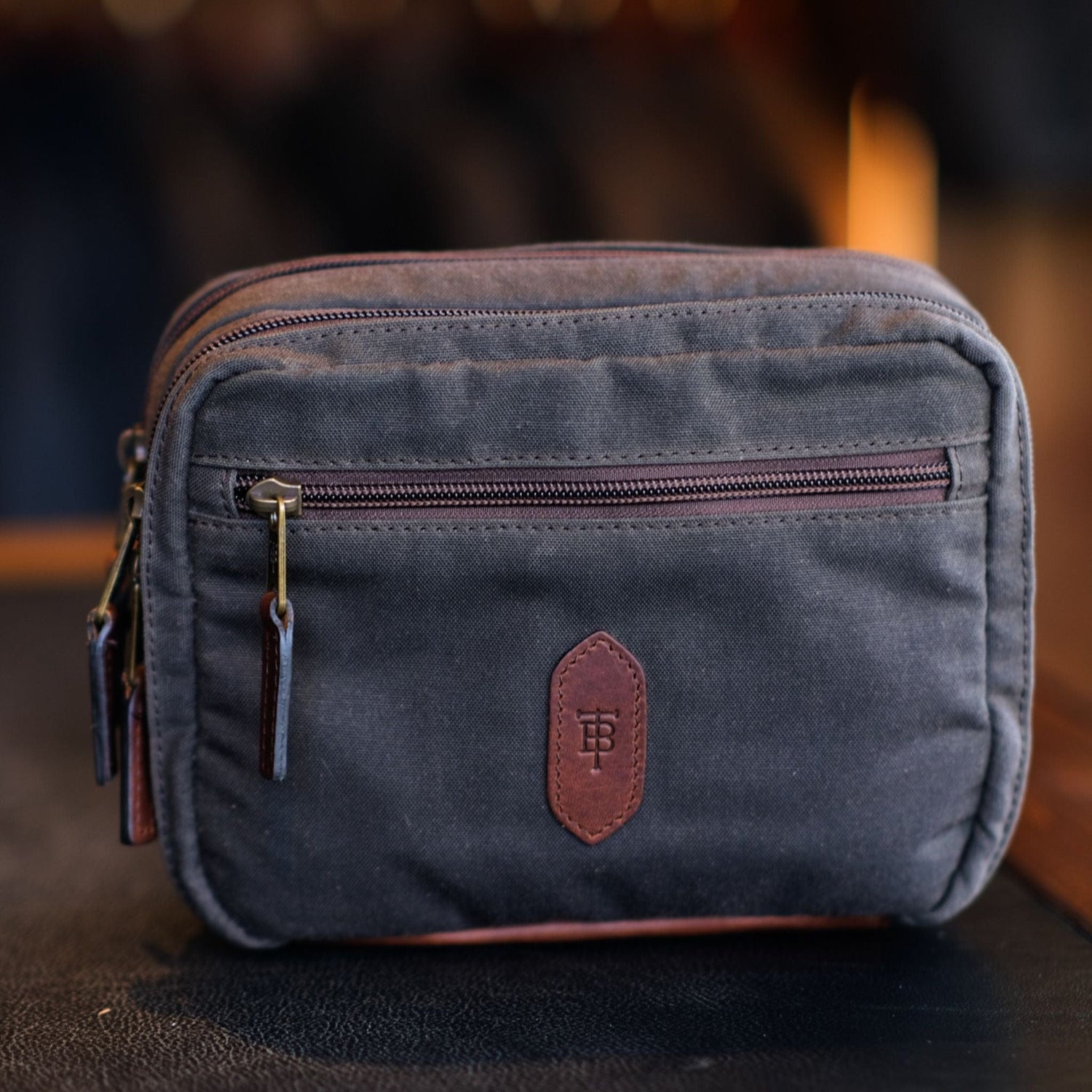 A Tom Beckbe dark gray Canvas Hanging Toiletry Bag with leather trim and a front zip pocket on a blurred background.