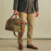 A person in khaki pants and brown shoes holds the Tom Beckbe Canvas Weekender Duffel Bag.