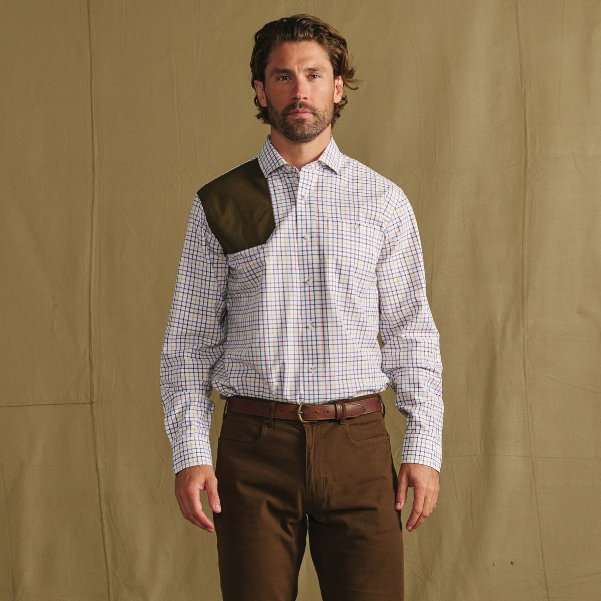 Man stands against a beige backdrop wearing a plaid shirt with a green shoulder patch and brown pants.