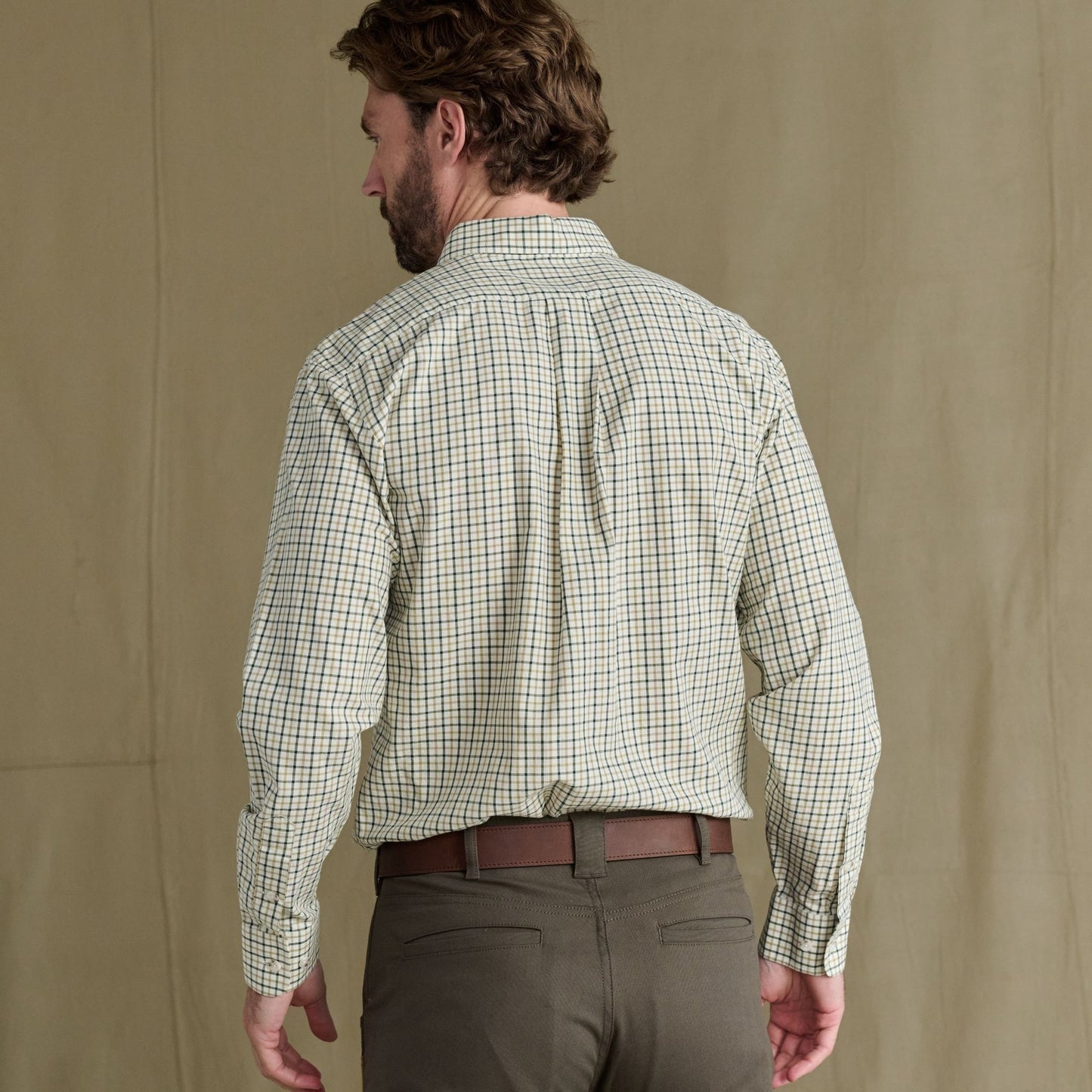 Man with brown hair wearing a light plaid shirt and dark pants, seen from behind, standing against a plain background.
