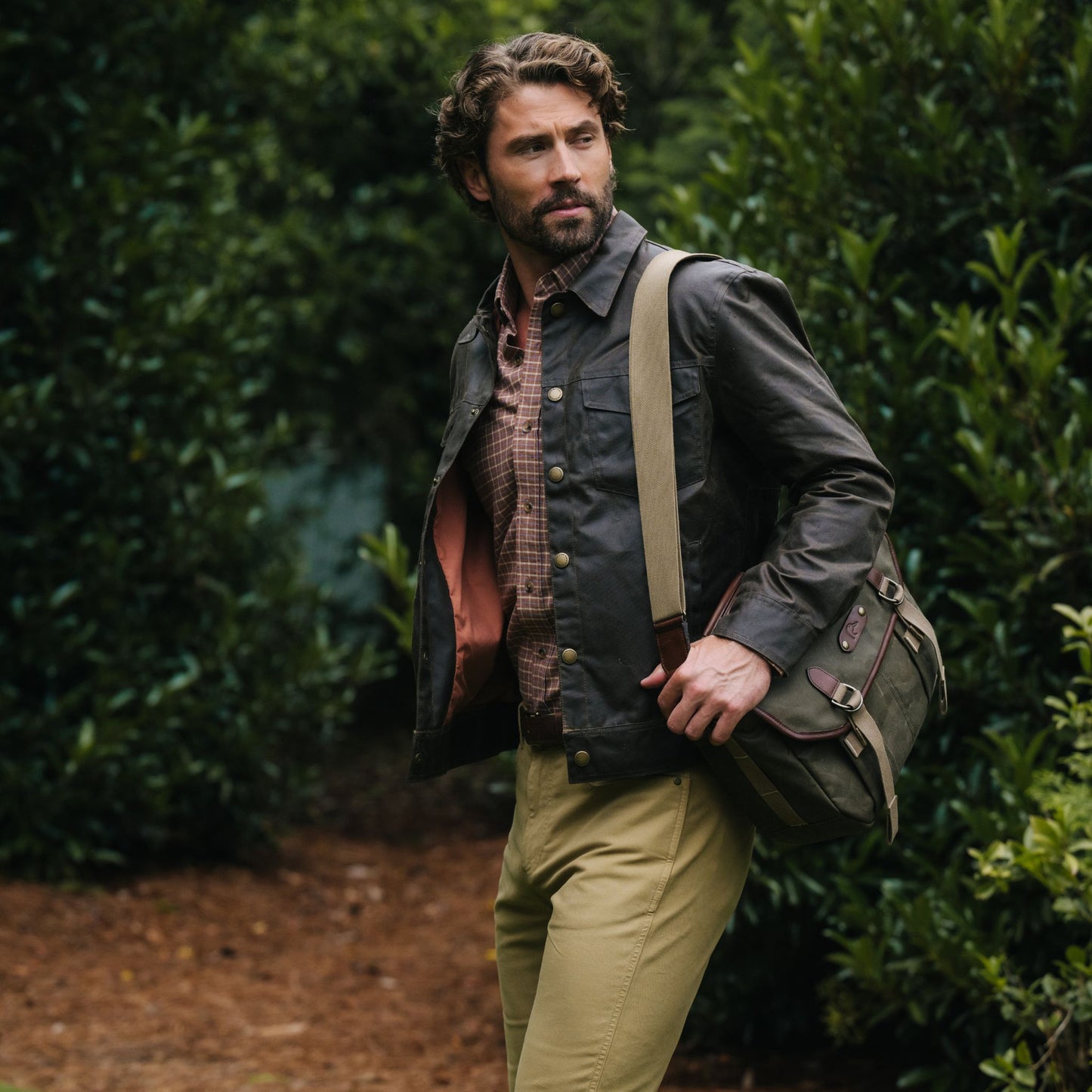 Bearded man in Tom Beckbe Chatom Jacket, plaid shirt, and khakis carries a shoulder bag in a wooded setting.