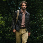 A man in a Tom Beckbe Chatom Jacket, plaid shirt, and khakis strolls outdoors with a shoulder bag among greenery.