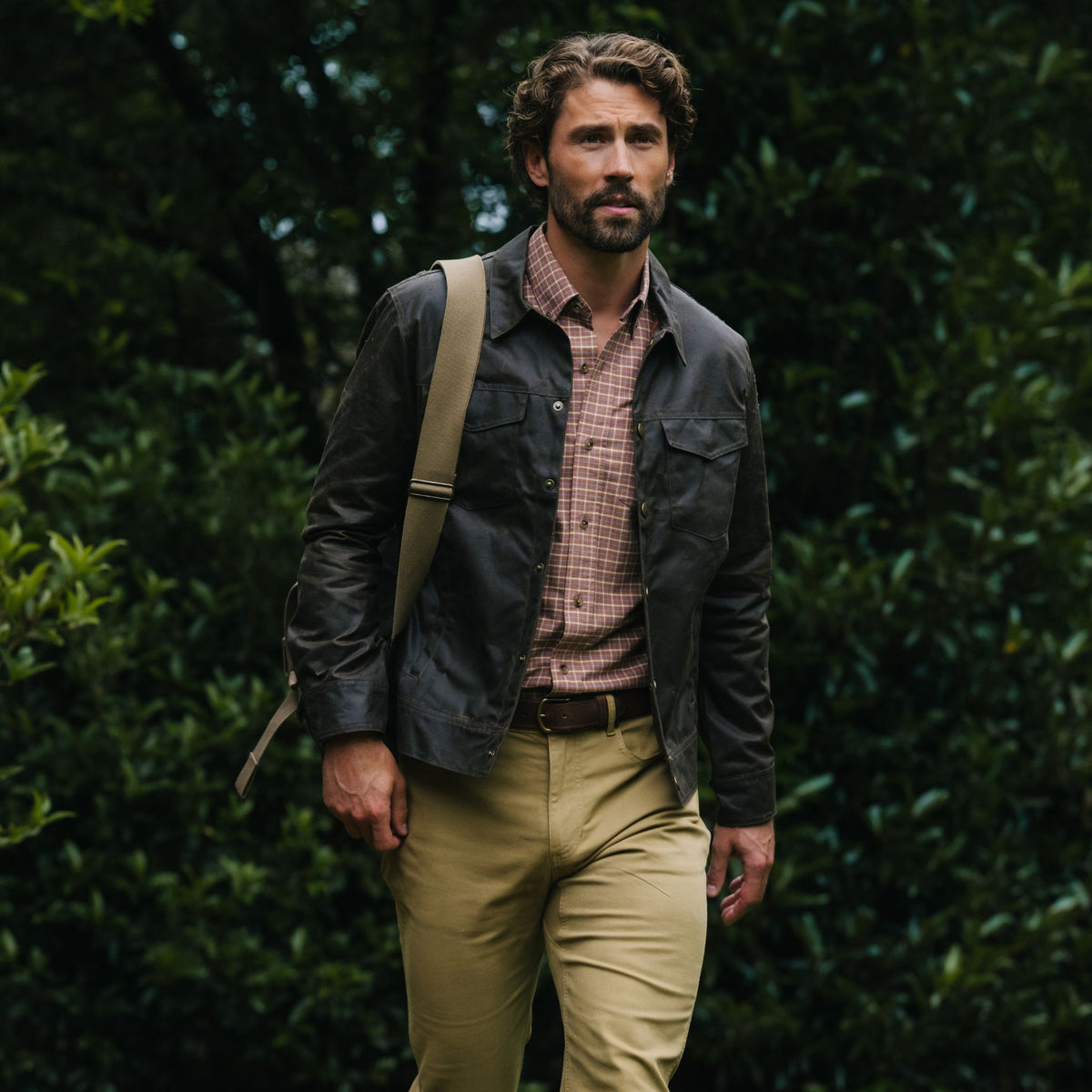A man in a Tom Beckbe Chatom Jacket, plaid shirt, and khakis strolls outdoors with a shoulder bag among greenery.