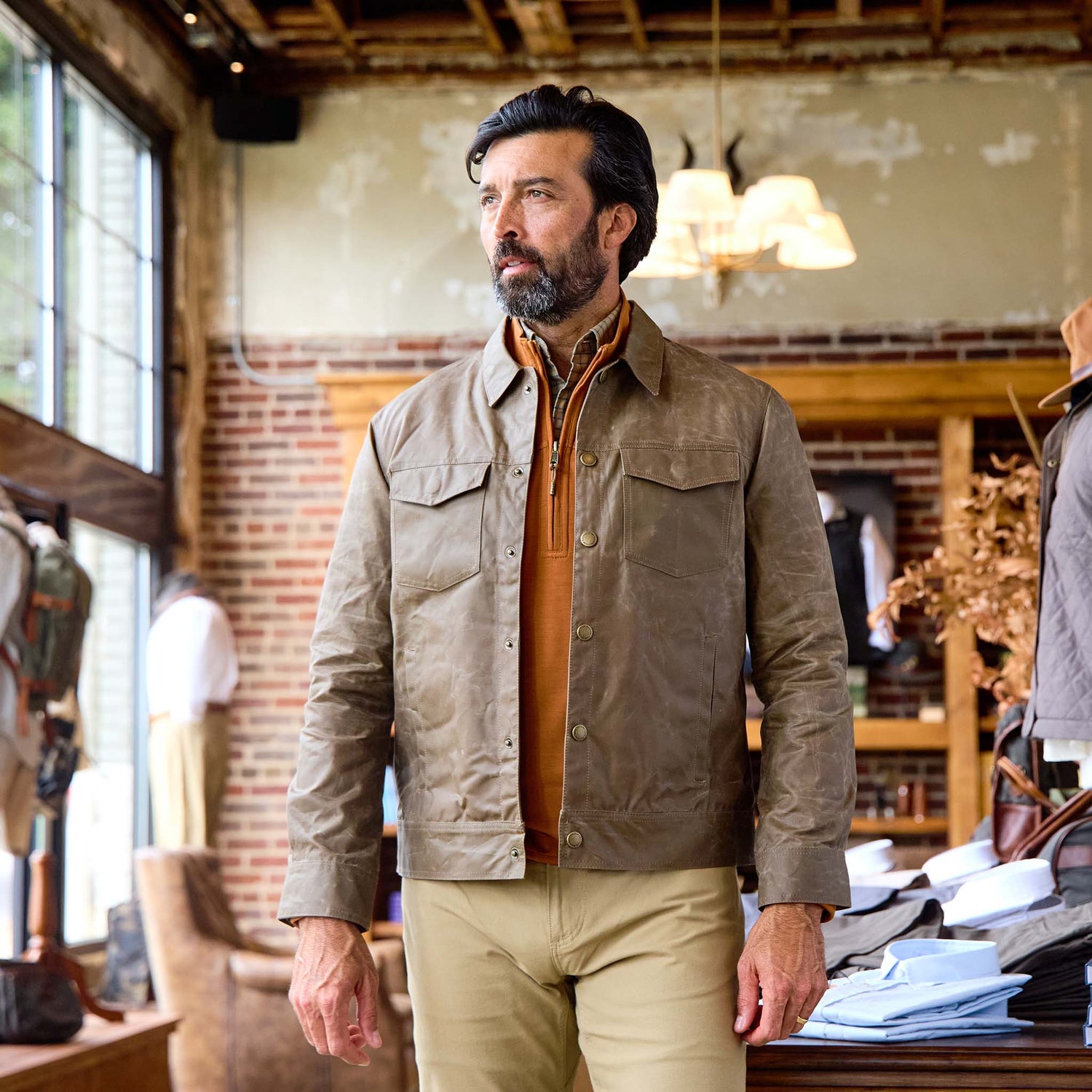 A man with dark hair and a beard wears a Tom Beckbe Chatom Jacket in a clothing store with brick walls and large windows.