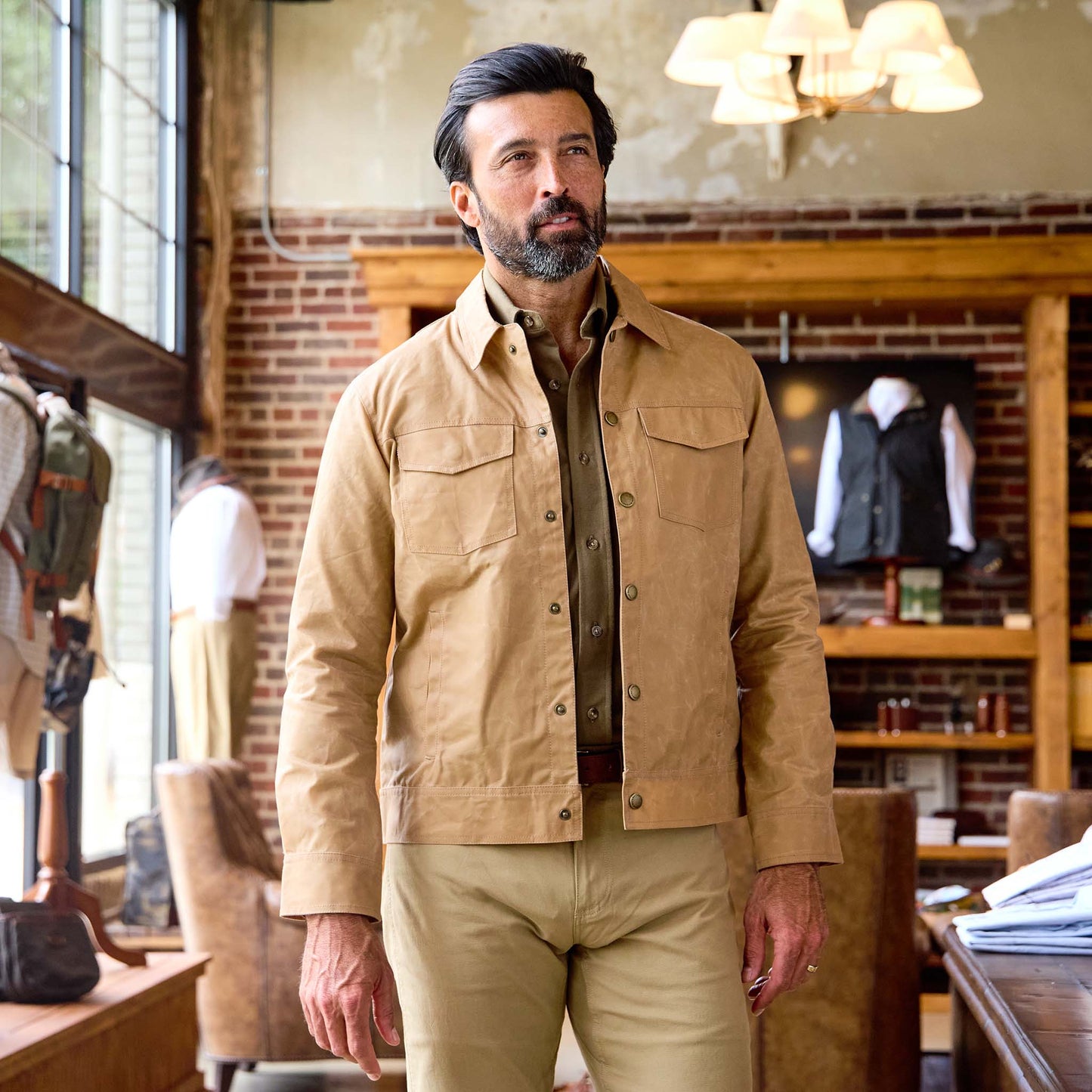 A man in a Tom Beckbe Chatom Jacket and khaki pants stands in a stylish room with brick walls and large windows.