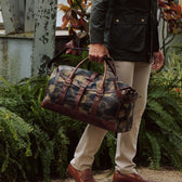 A person in khaki pants holds a Tom Beckbe Classic Camo Weekender Duffel Bag with brown leather accents by green plants.