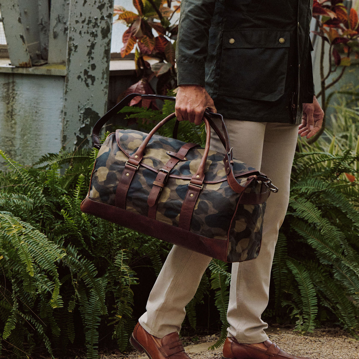 A person in khaki pants holds a Tom Beckbe Classic Camo Weekender Duffel Bag with brown leather accents by green plants.