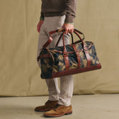Man in beige pants holds a Tom Beckbe Classic Camo Weekender Duffel Bag with brown leather accents on a neutral background.