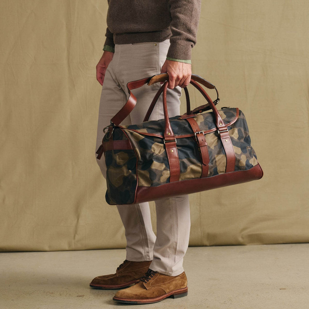 Man in beige pants holds a Tom Beckbe Classic Camo Weekender Duffel Bag with brown leather accents on a neutral background.