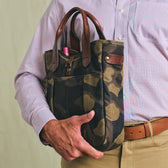 A person holds the Tom Beckbe Classic Camo Whiskey Tote with brown leather handles and a bottle peeking out.