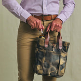 A person puts a beer into the Tom Beckbe Classic Camo Whiskey Tote while wearing khaki pants and a belt.