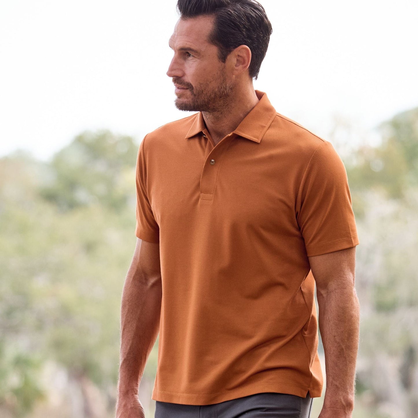 A man in a Tom Beckbe Coastal Polo stands outdoors gazing sideways, trees and the clear sky in the background.