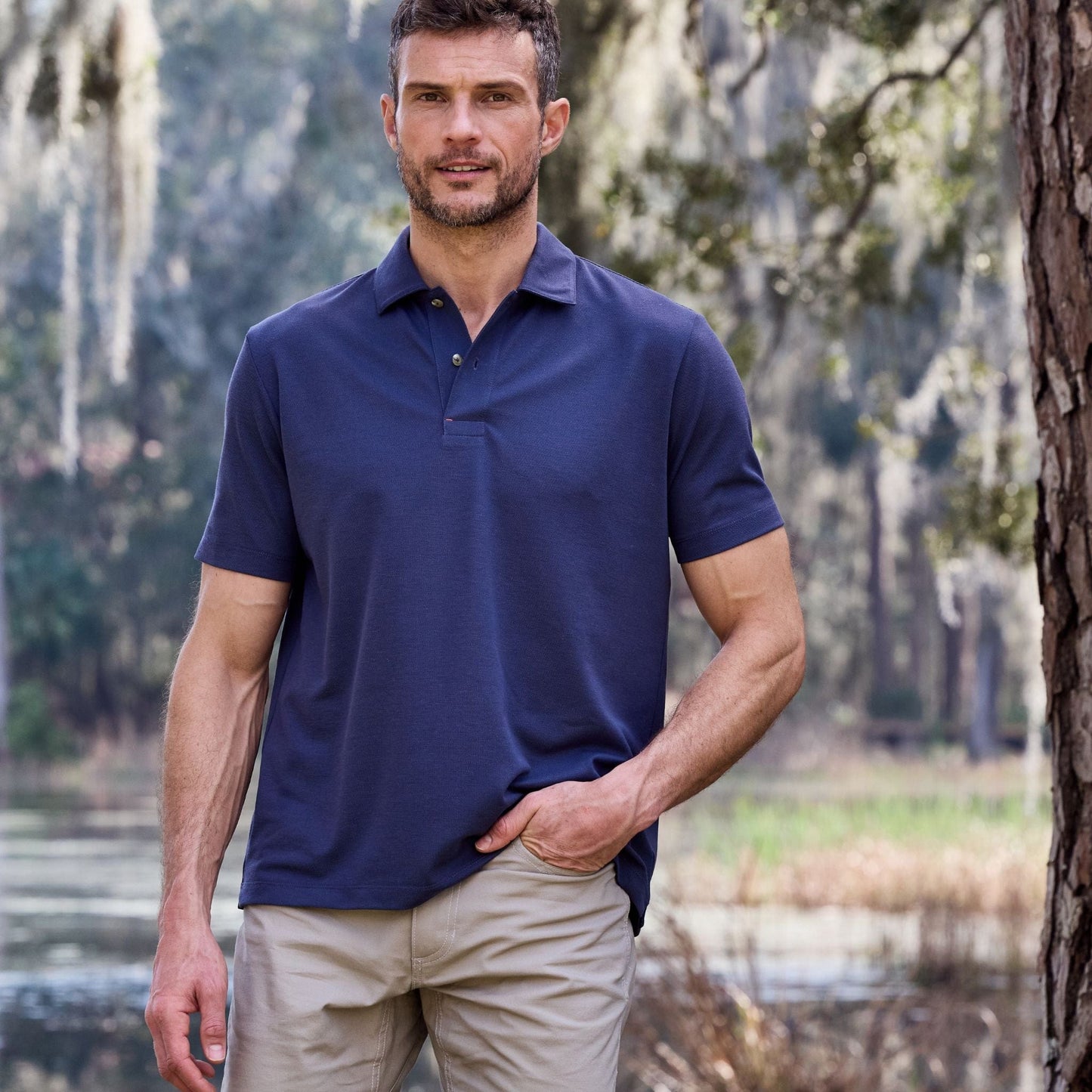 A person in a navy Tom Beckbe Coastal Polo Shirt and beige pants stands outdoors near trees, a pond, and moss.