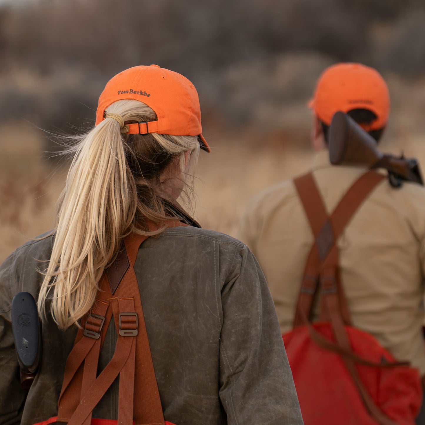 Two people in Tom Beckbe Cotton Twill Baseball Hats and hunting gear walk through tall grass, shotgun over shoulder.
