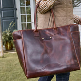 Person carrying a large brown leather tote bag outdoors near a house with glass doors and greenery.