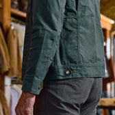 A man wearing the Tom Beckbe Crawford Jacket and gray pants stands indoors with clothing racks behind him.